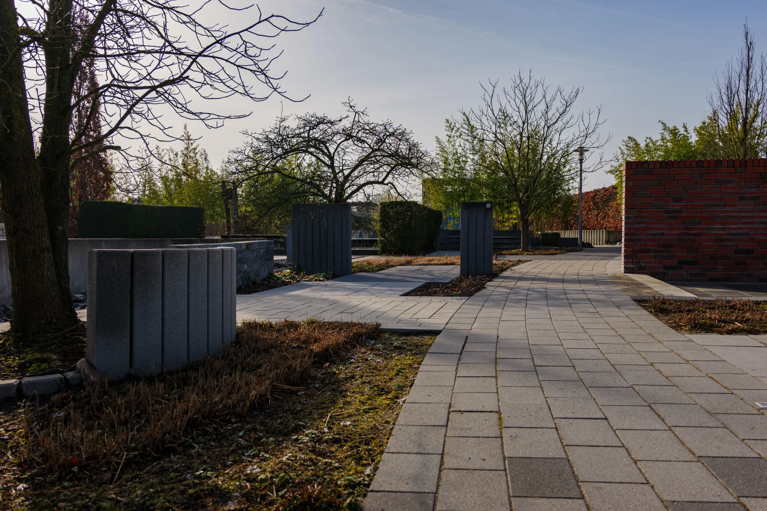 Park walkway with gray pavers, leafless trees, and a red brick wall along the right under a clear blue sky.