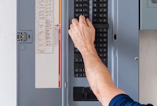 Close-up of a person flipping a circuit breaker in a panel, with a hand on a row of black breakers and the panel door open showing labeling on the side.
