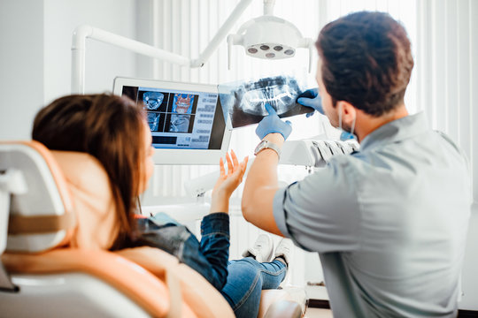 Dentist showing dental X-ray images to patient in a treatment room with a monitor displaying scans.